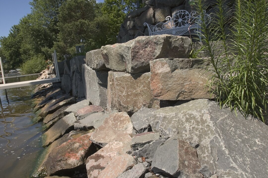 Lakeside stone wall with bench and greenery.