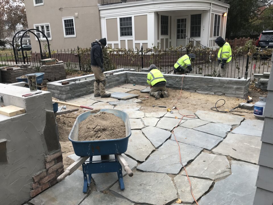 Workers laying stone patio with wheelbarrow nearby.