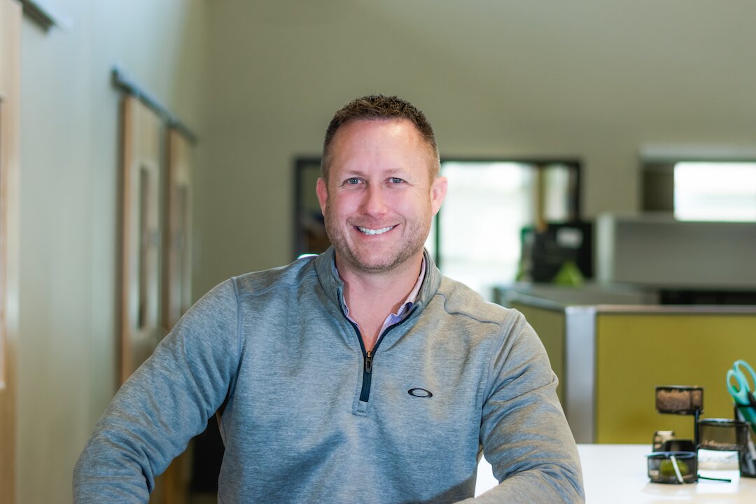 Smiling man in office setting, gray sweater.