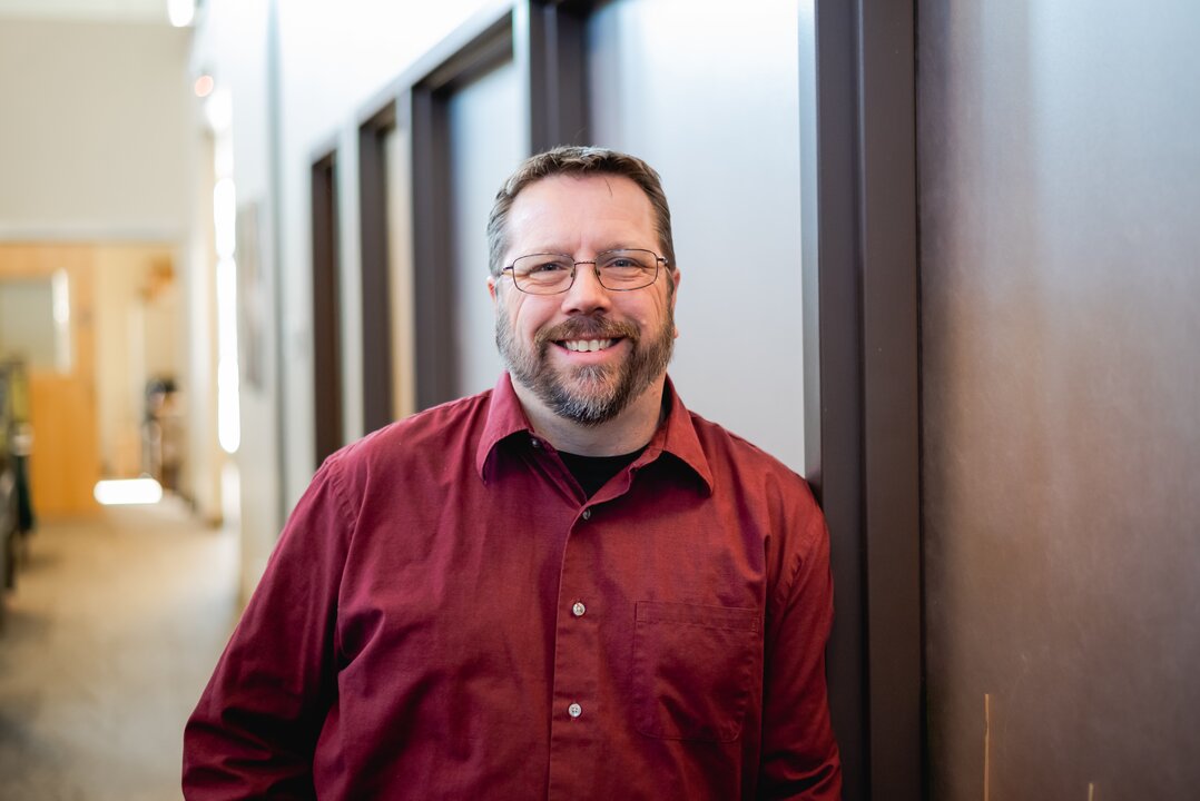 Man smiling in office hallway, wearing red shirt.