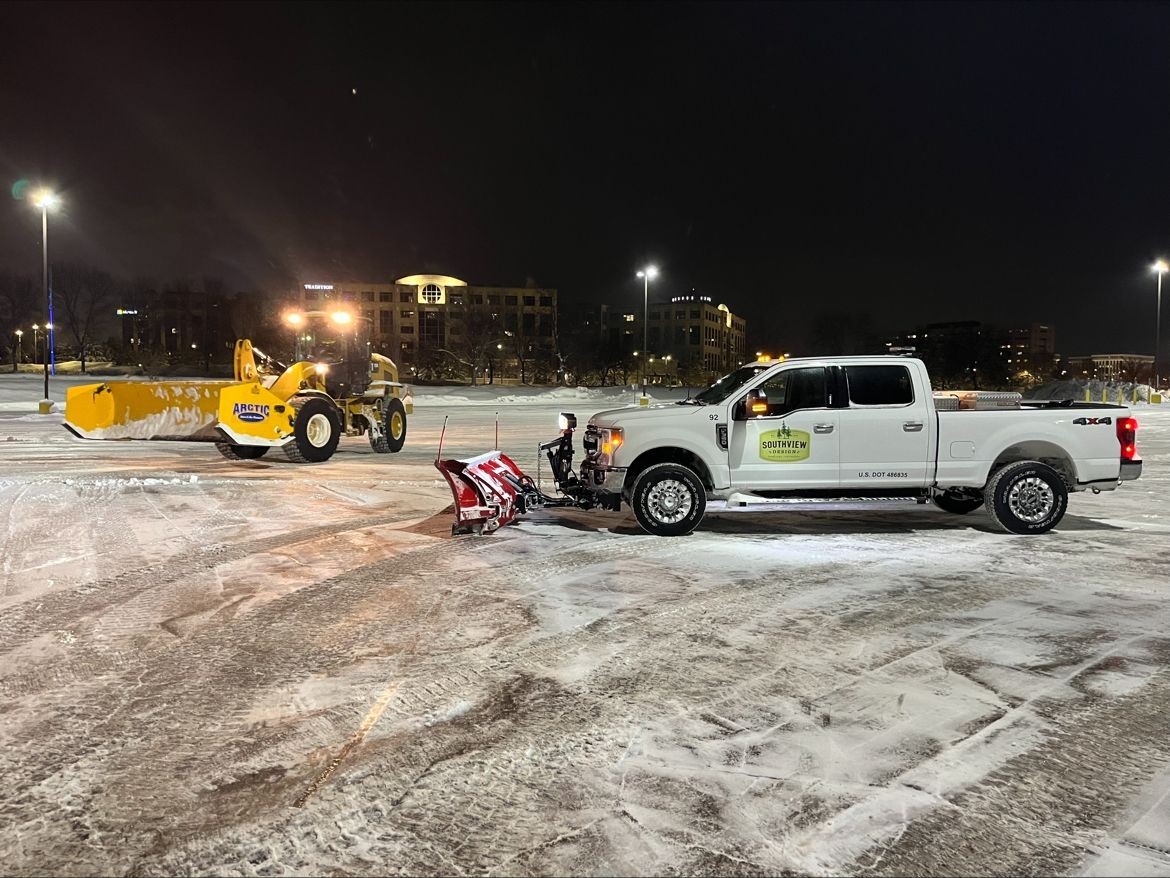 Snow plow truck clearing snowy parking lot at night.