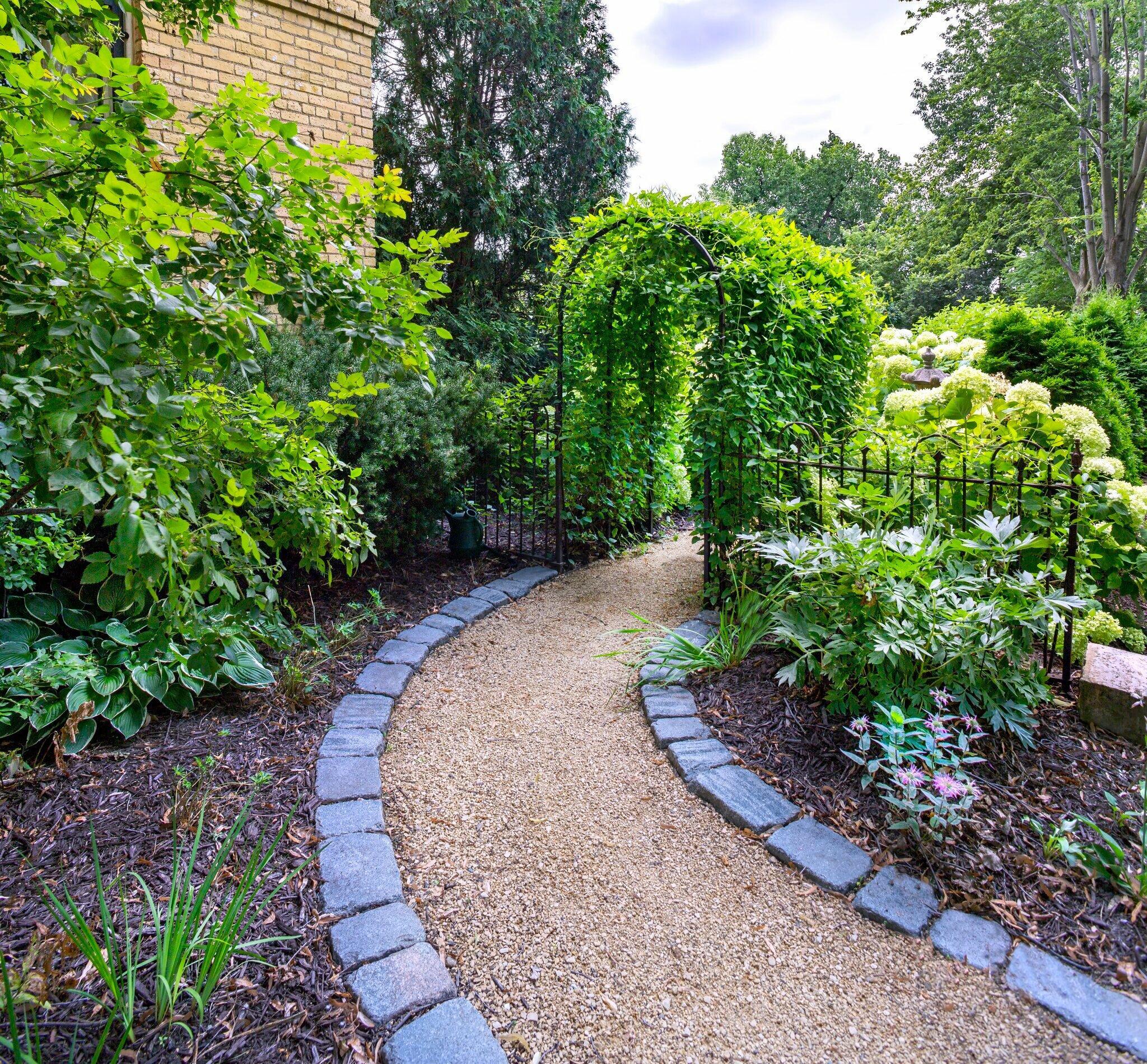 A charming garden pathway lined with perennials and framed by a lush vine-covered arbor — a perfect example of immersive, nature-forward landscape design.