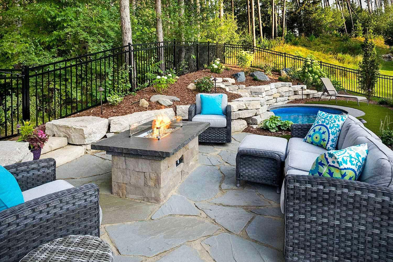 A cozy stone patio with modern fire table, layered retaining walls, and lush plantings — blending comfort, functionality, and natural materials.