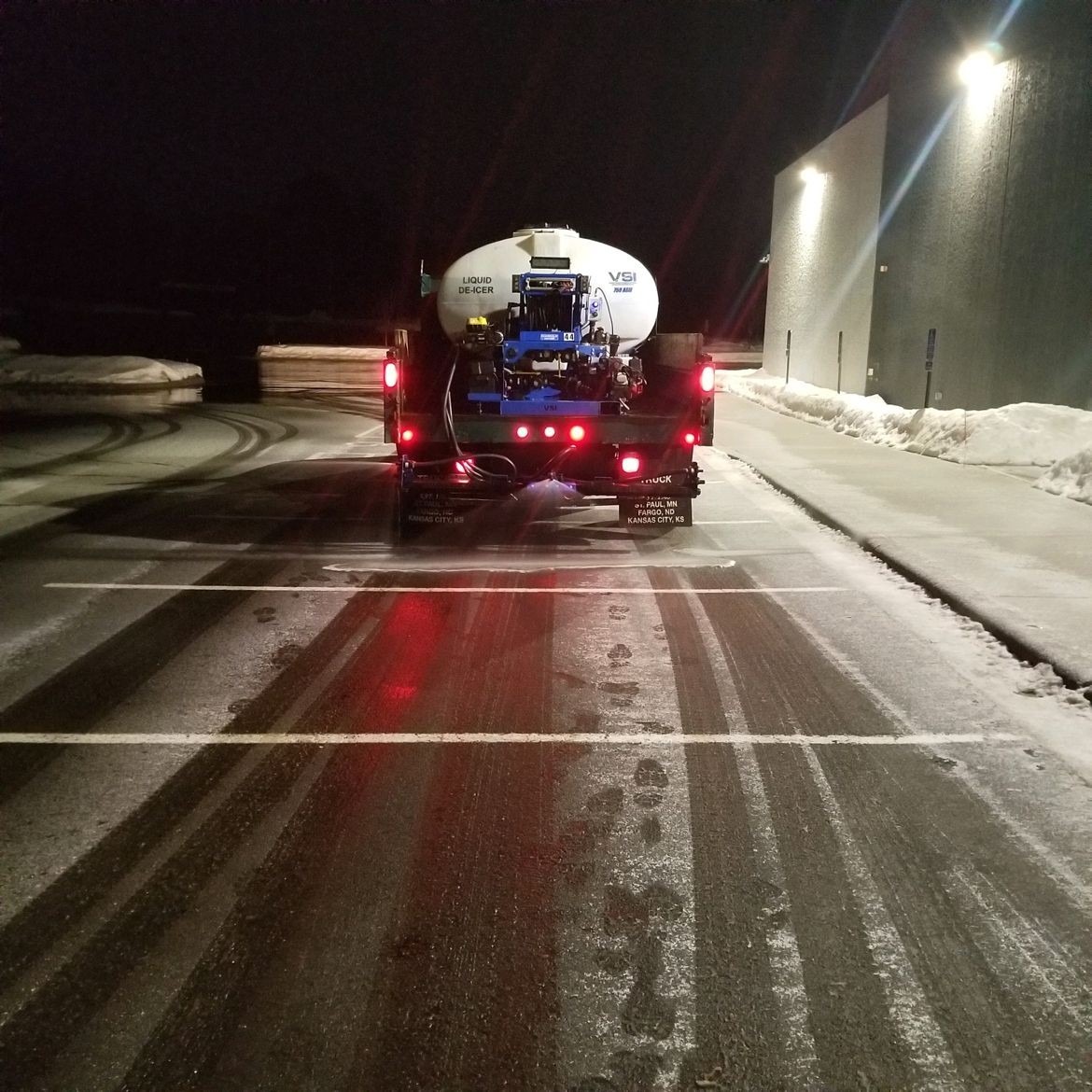 Truck applying de-icer on snowy parking lot at night.