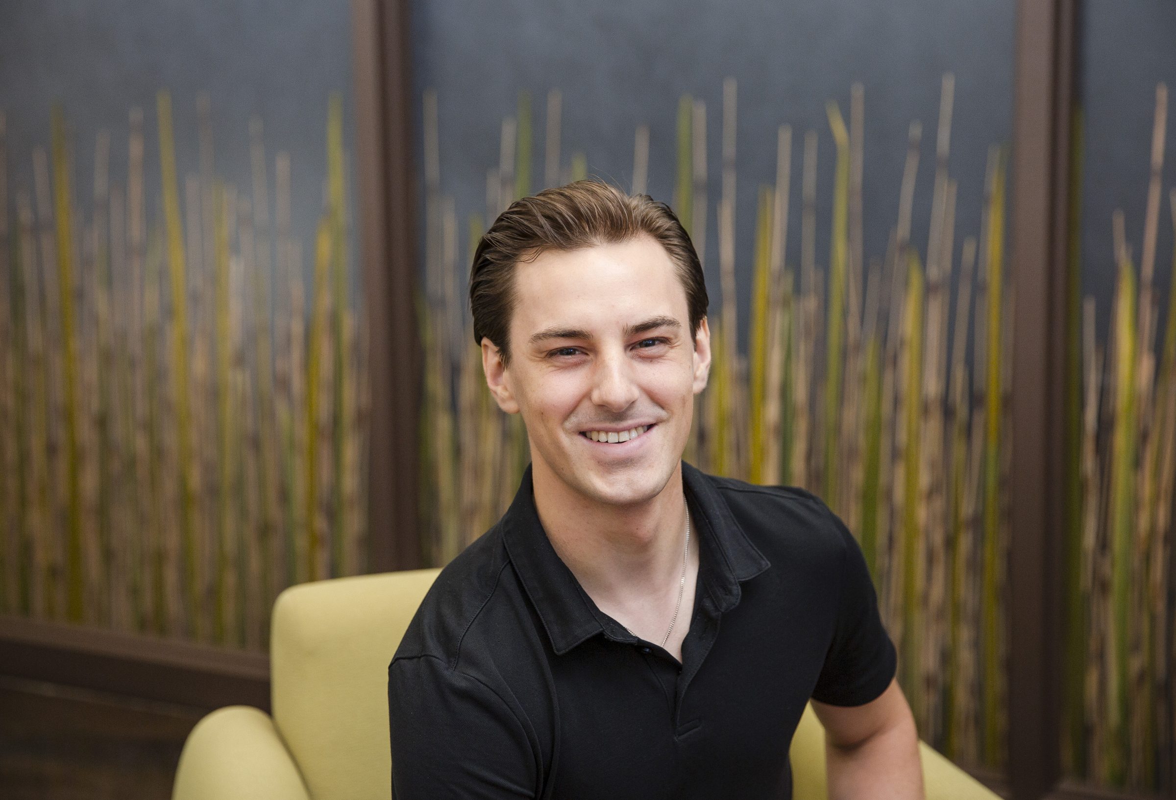 Smiling person seated indoors with bamboo backdrop.