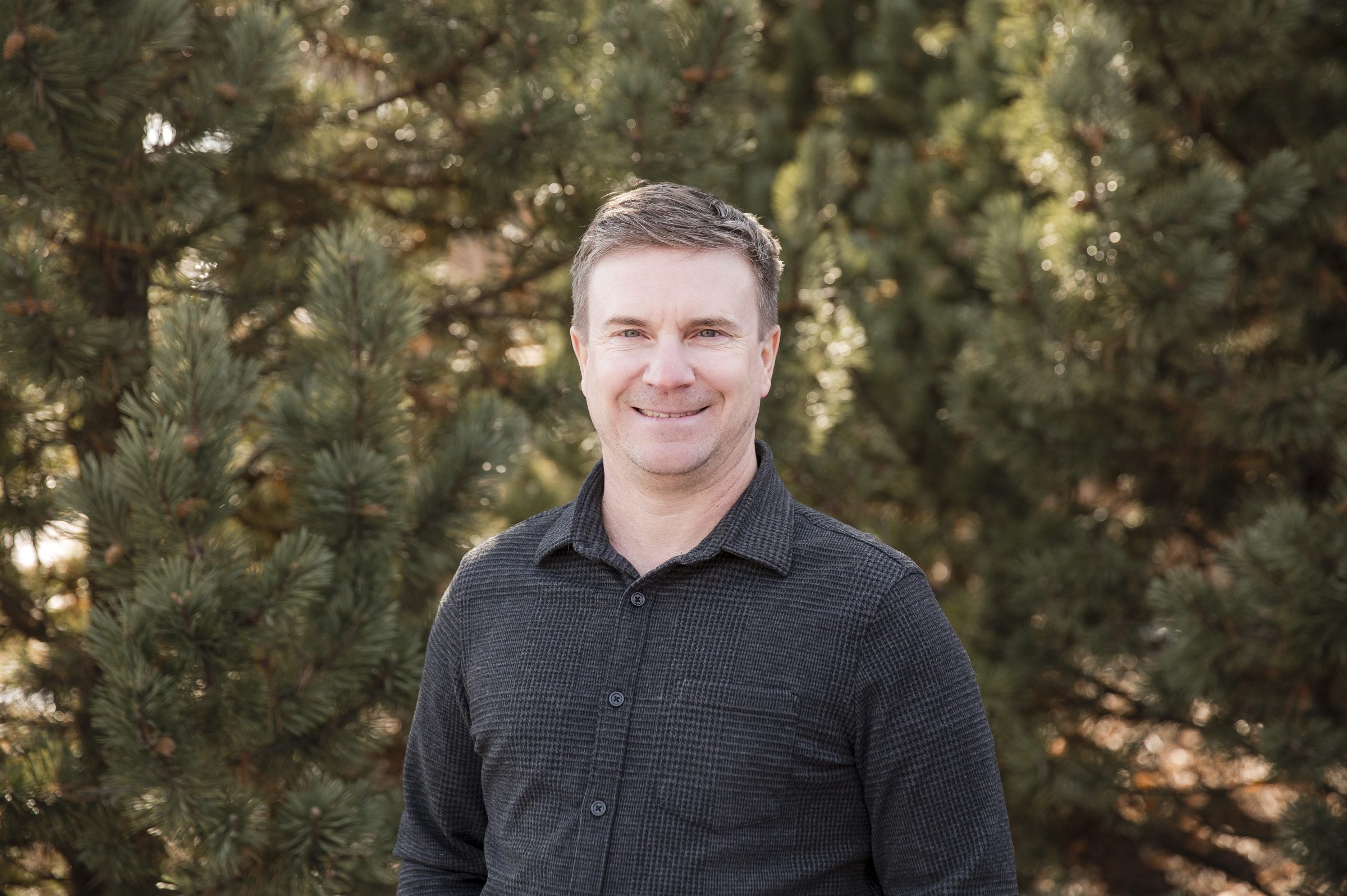 Smiling man standing near green pine trees.