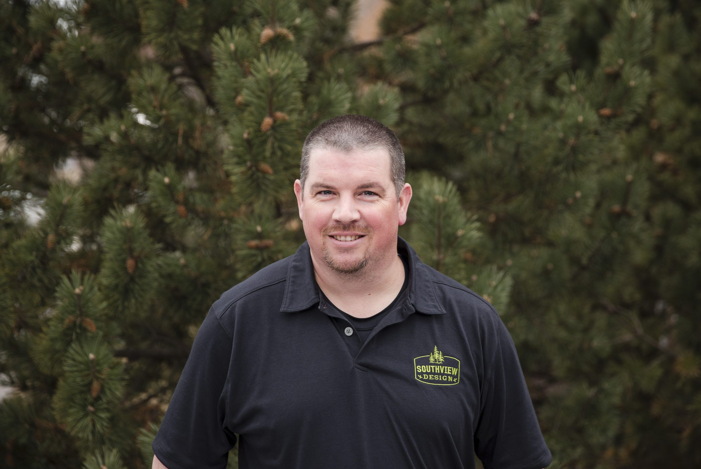 Man in black polo shirt, outdoors with trees.