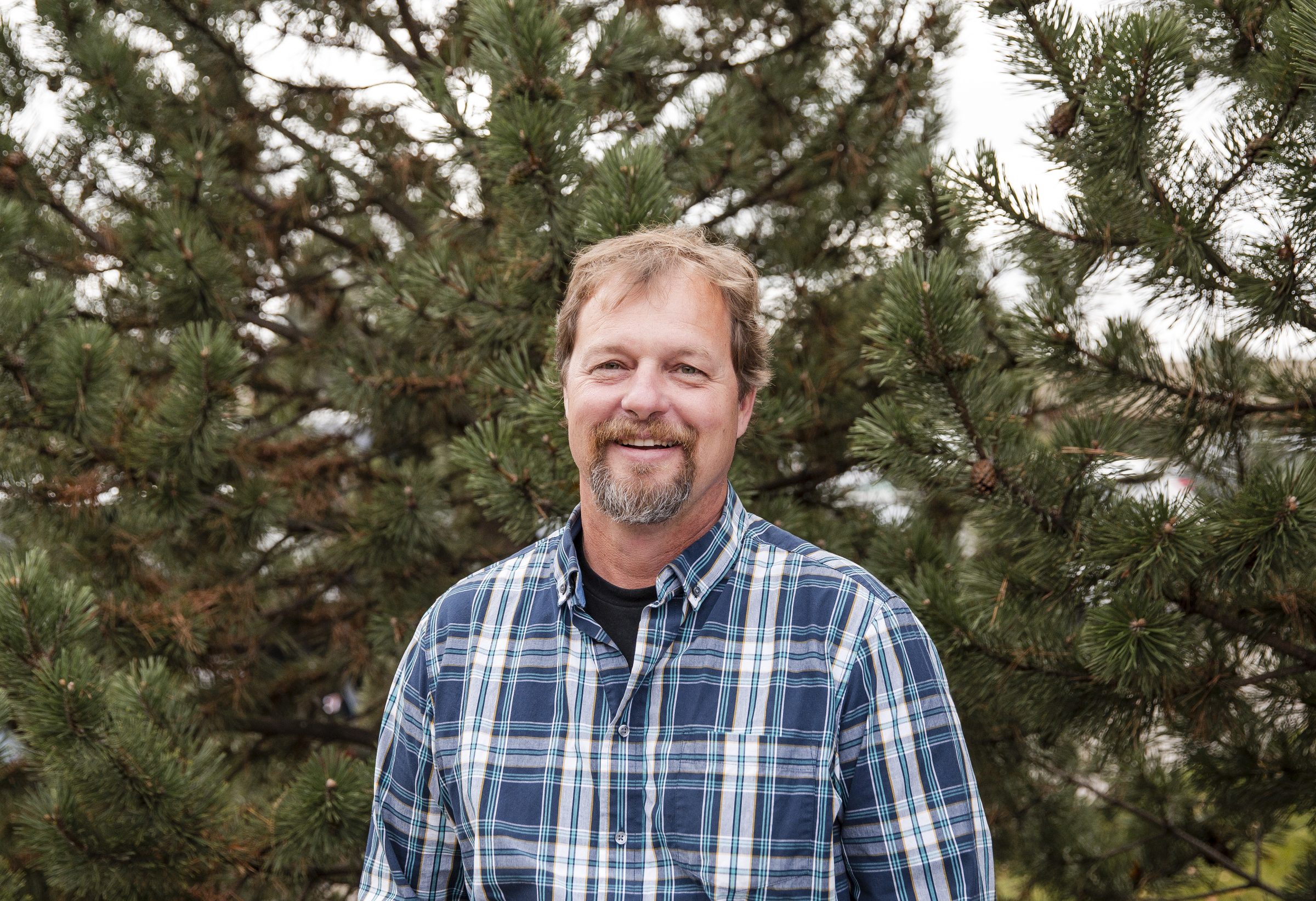 Smiling man in plaid shirt among pine trees.