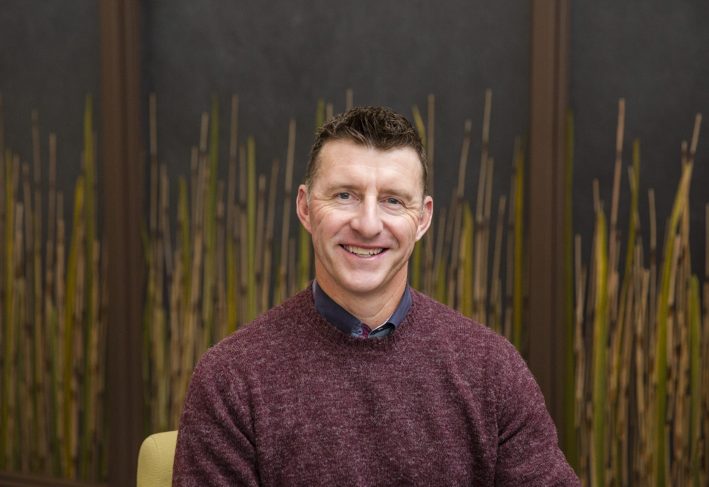 Smiling man in brown sweater with bamboo background.