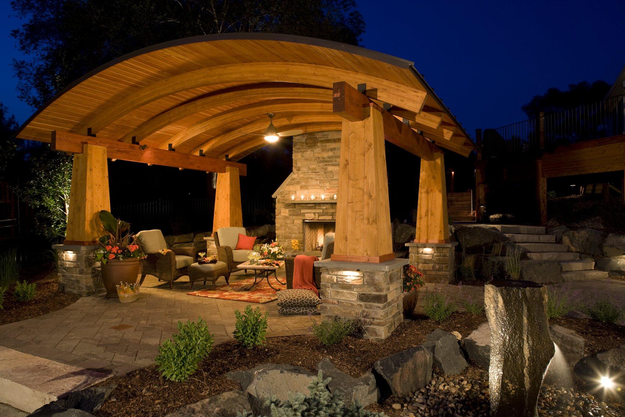 Curved wood beam pavilion with stone fireplace and paver patio lit up at night