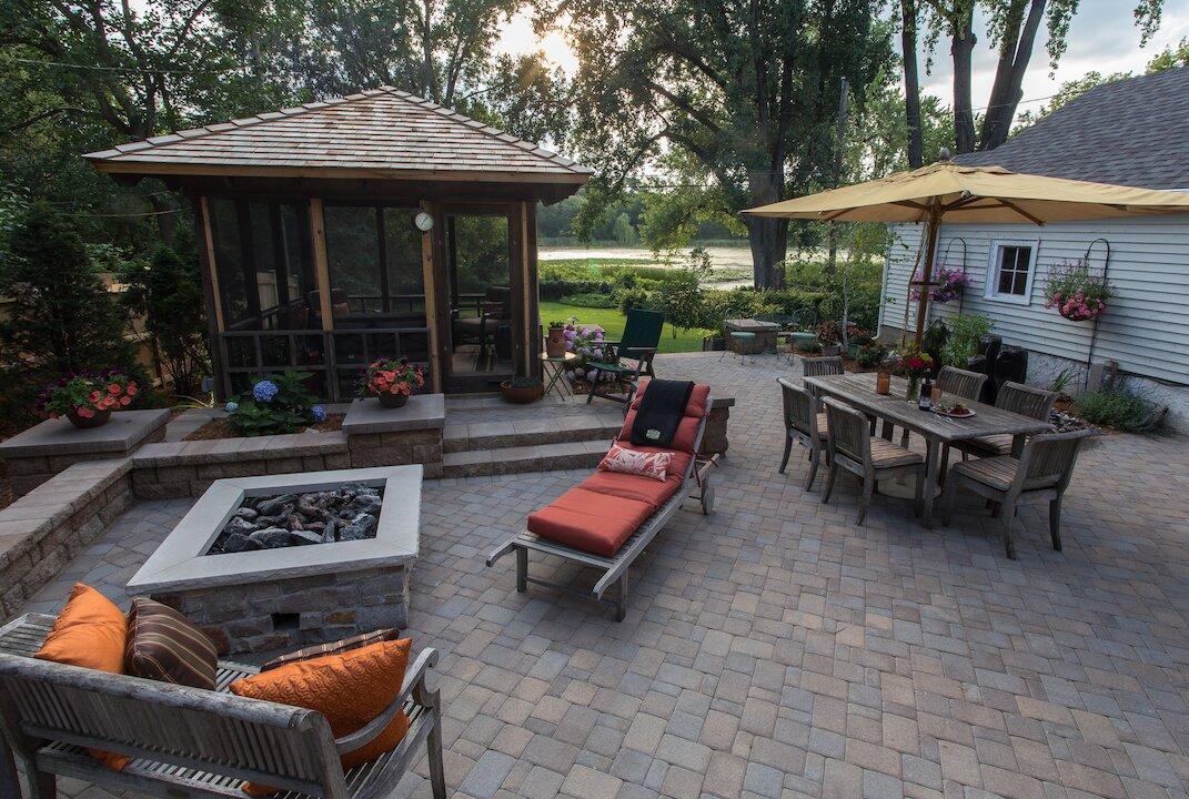Large paver patio with fire pit, dining area, and screened gazebo overlooking a lake