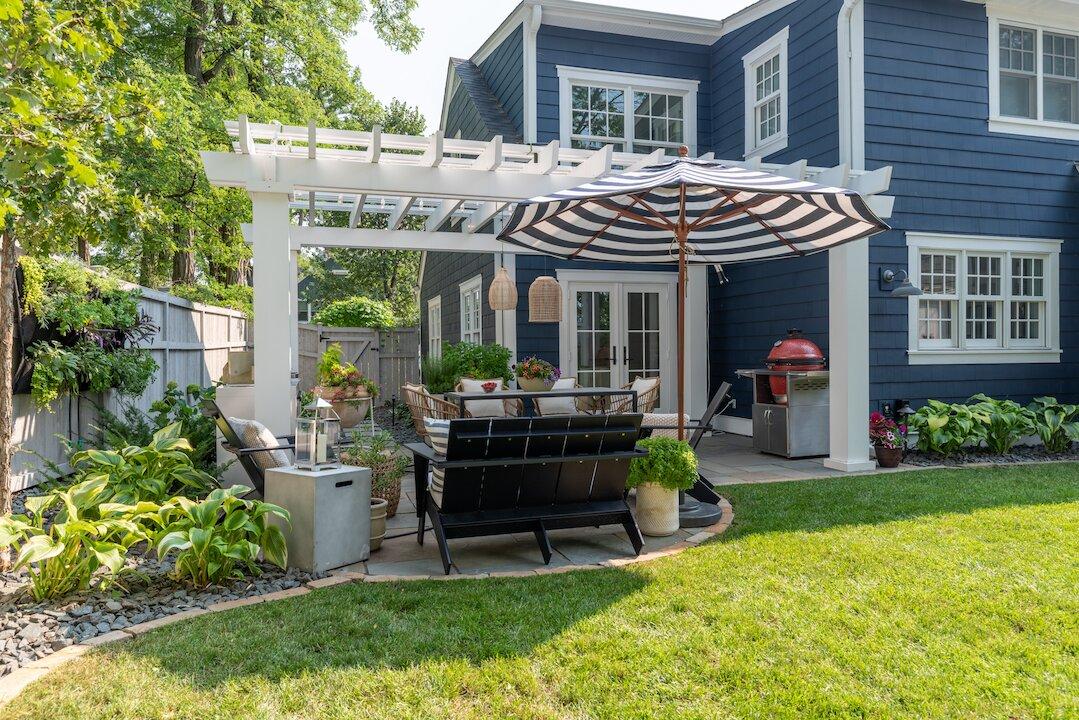 White pergola over stone patio with outdoor dining and lush garden beds