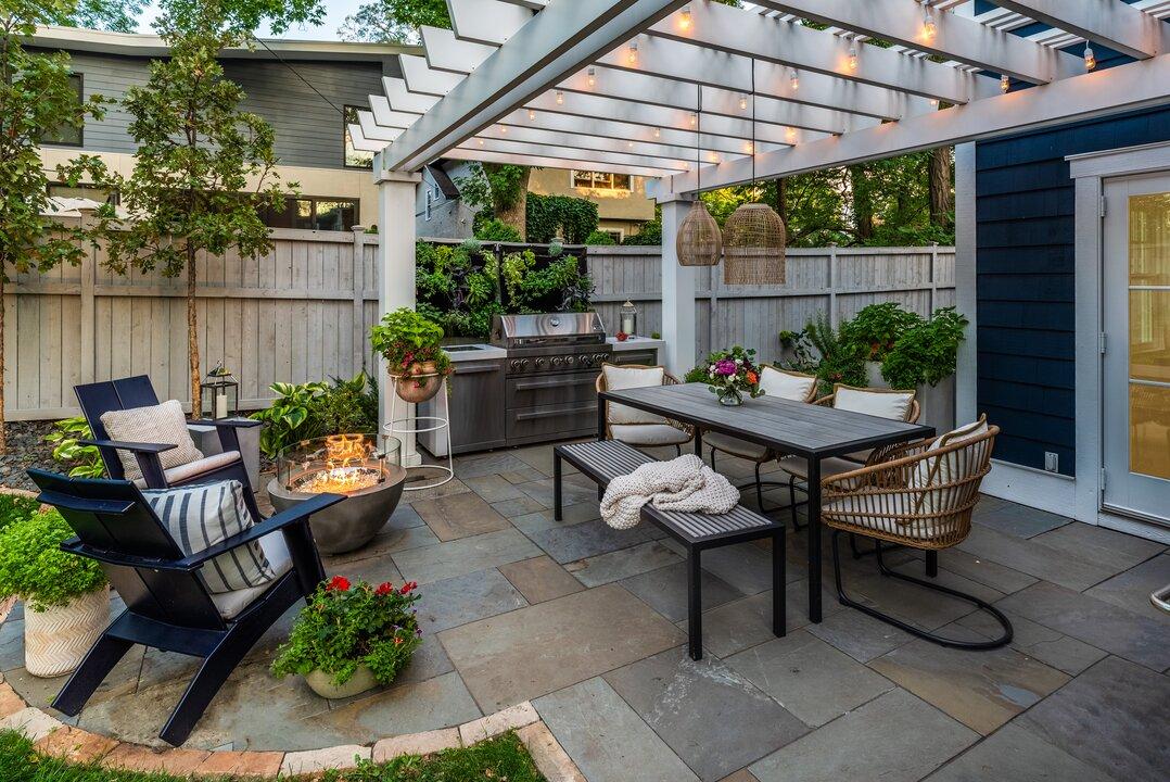 White pergola with string lights over flagstone patio with fire pit and outdoor kitchen