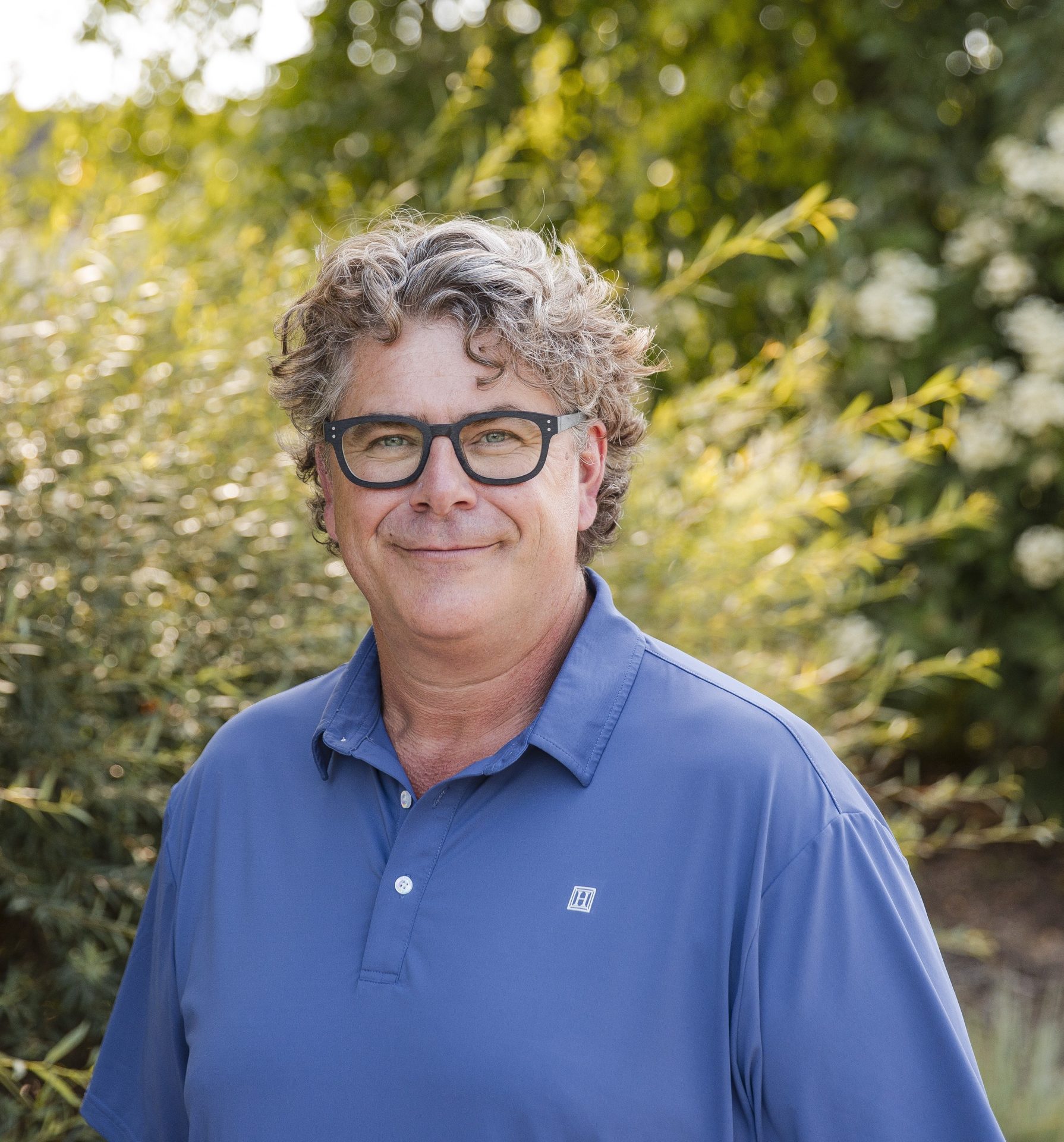 Man smiling outdoors in blue shirt and glasses.